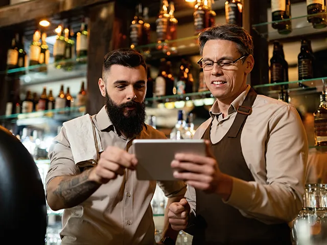 Zwei Männer stehen in einer Bar und besprechen etwas an einem Tablet.
